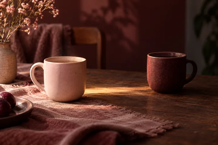 A light pink cup and a dark red cup sit apart on a wooden table, with flowers and a plate of dark fruit nearby.