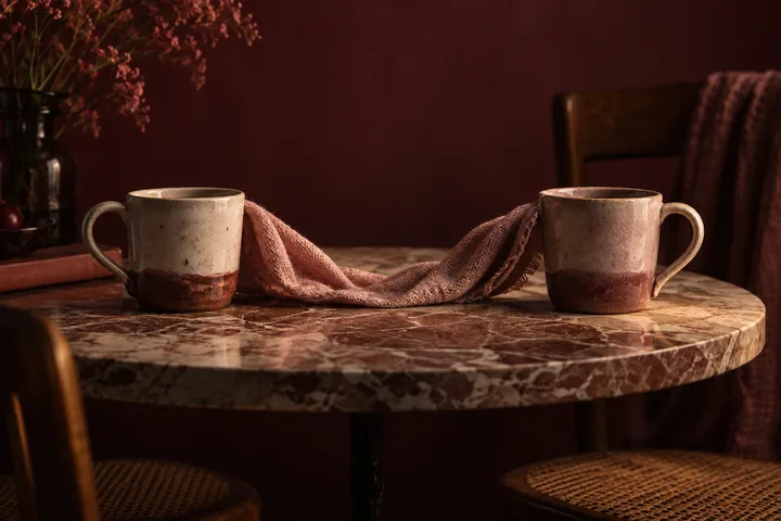 Two ceramic mugs sit apart on a marble table with a cloth draped between them.