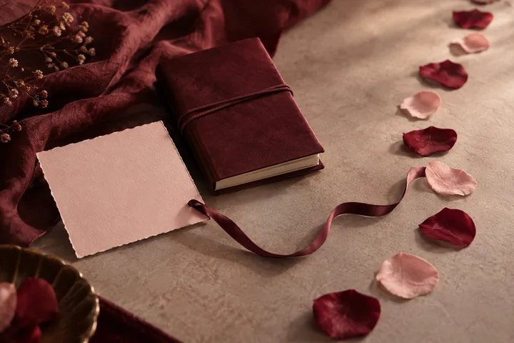 A closed burgundy book rests beside a blank sheet of paper and a trailing burgundy ribbon, surrounded by scattered rose petals.