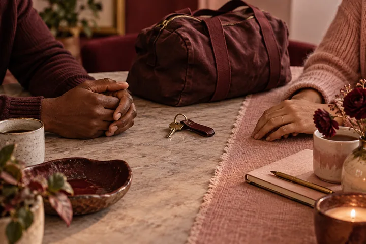 Two people sit across a table from a burgundy overnight bag and a set of keys, a warm editorial scene about discussing practical help without handing over a woman’s home or money.