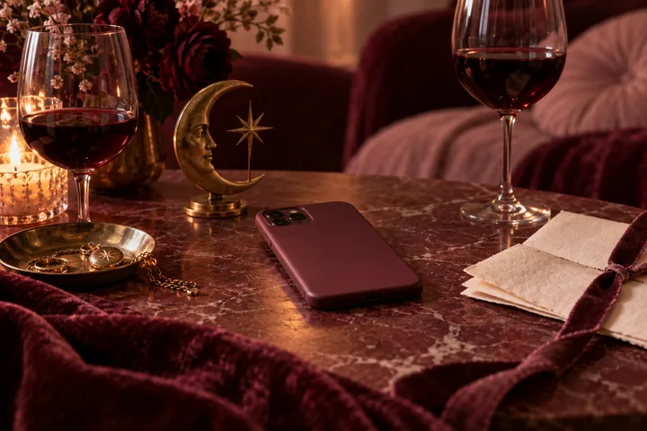 A burgundy phone and open notebook sit on a marble table between two glasses of red wine, beside a crescent moon ornament and candle.