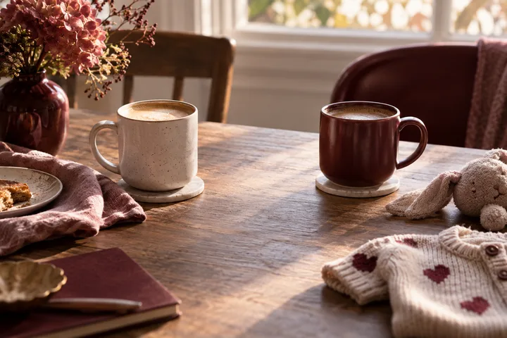 Two mugs sit on a wooden table beside a small heart-patterned sweater, plush rabbit, flowers and folded fabric.