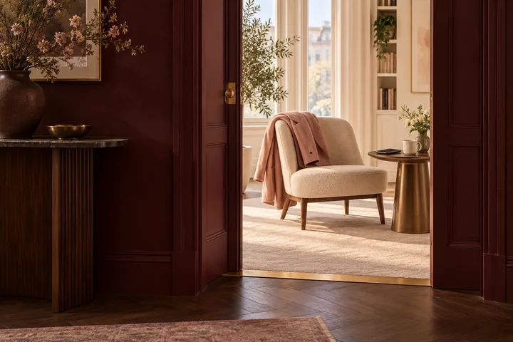 A burgundy doorway opens onto a warm room with a cream chair, blush coat, and brass threshold line symbolizing a calm boundary.