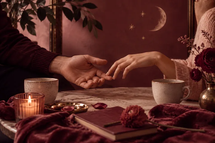 Two hands reach toward each other across a table set with mugs, a candle, flowers, and a closed book; a crescent moon and stars decorate the burgundy wall.