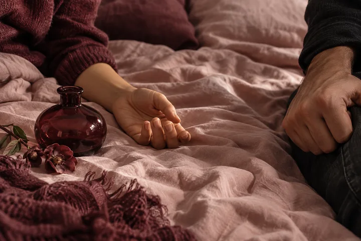 Two visible arms and hands rest on a rumpled pink bedspread beside a dark glass bottle and flowers.