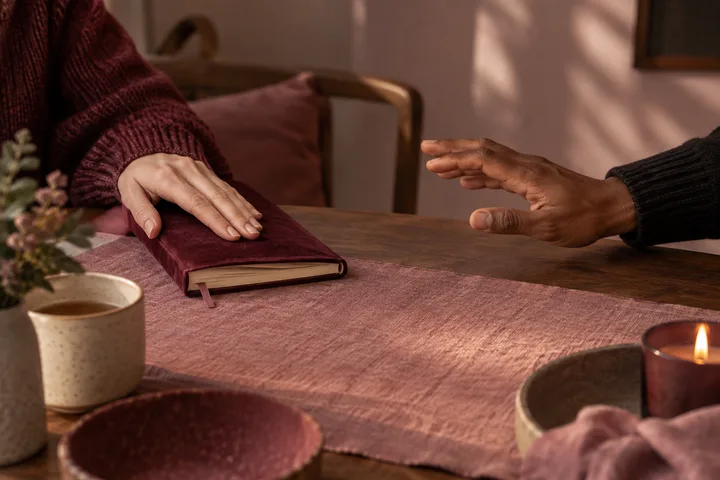 Two hands reach toward each other across a wooden table with a burgundy book between them; mugs and a lit candle sit nearby.