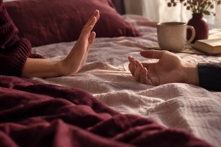 Two hands are shown on a bed, with one palm raised and the other hand open; a mug, books, and a vase are in the background.