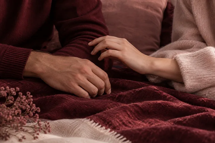 A woman's hand rests gently on a man's sleeve beneath a burgundy blanket, suggesting quiet physical reassurance.