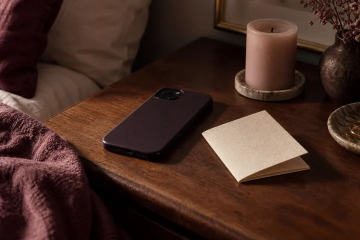 A phone and folded handwritten note sit on a wooden bedside table beside a lit blush candle, suggesting a pause before a reply.