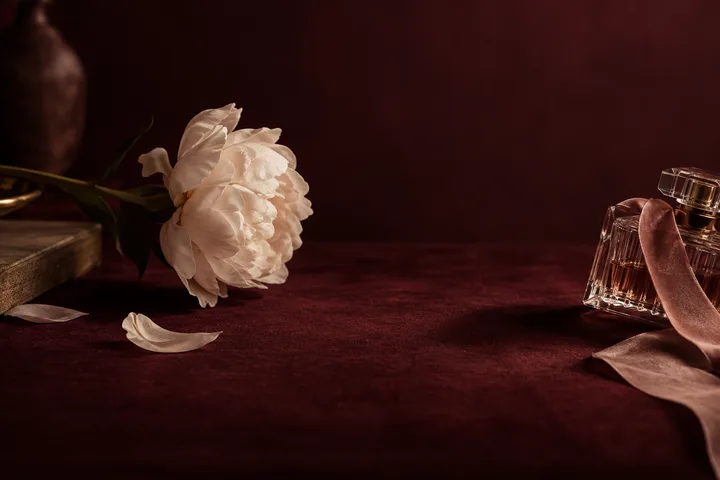 White flower resting beside a glass perfume bottle on burgundy fabric, a still life for the contrast between appealing attention and dependable care.