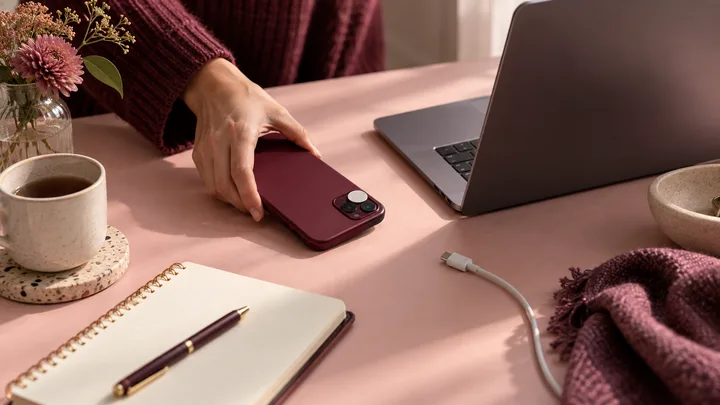 A hand holds a burgundy smartphone on a pink desk beside an open laptop, tea cup, notebook, pen, flowers, and charging cable.