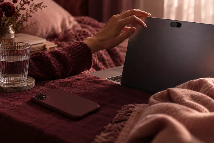 A hand reaches toward the camera on an open laptop, with a smartphone on a burgundy blanket and a glass of water nearby.