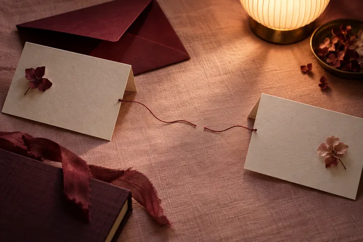 Two blank folded cards with small flowers are connected by loose red threads on a cloth-covered table. An open burgundy envelope, a dark book, a lamp, and a bowl of petals are nearby.