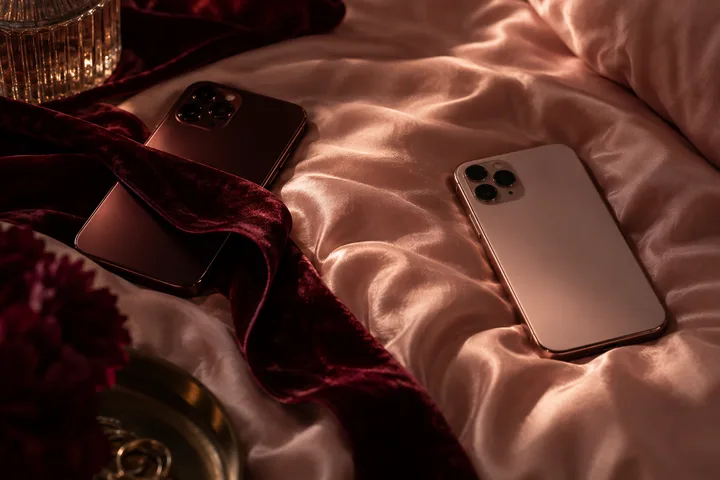 Two smartphones lie on pale pink bedding beside a burgundy velvet ribbon, flowers, and a glass.