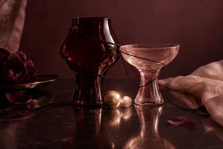 A dark burgundy glass vase and pale pink goblet are looped together with black cord on a reflective table, beside two small spheres, flower petals, and folded fabric.