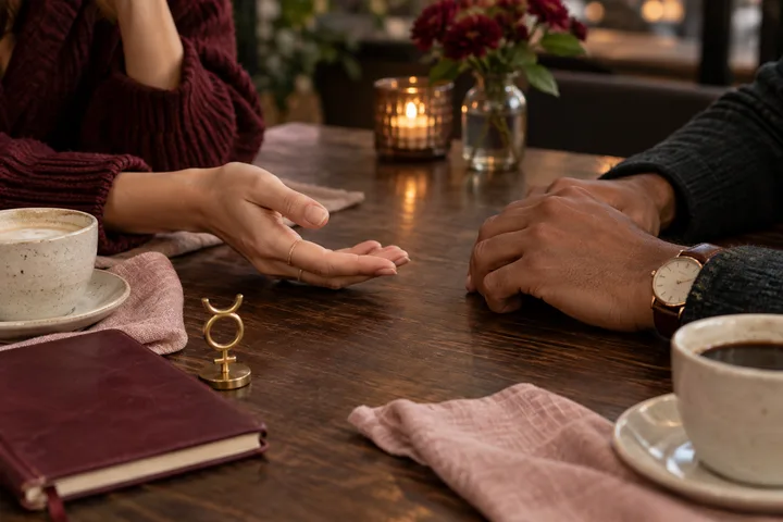 Two people’s hands pause across a café table, one hand making an open palm gesture, with cups, a burgundy notebook, and a brass Mercury symbol.