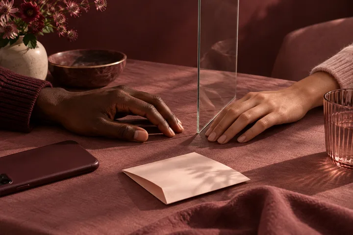 Two hands rest on opposite sides of a clear divider on a burgundy table, with a phone, blank envelope, flowers, and glasses nearby.
