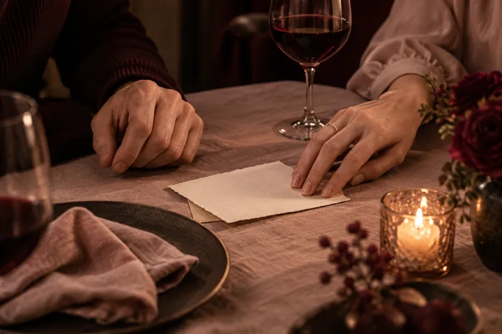 Two hands rest beside an unopened letter on a candlelit dinner table with wine and burgundy roses.