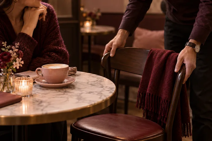 A person at a café table watches as another moves a chair draped with a burgundy scarf.