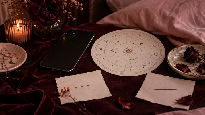 A black phone, zodiac chart, candle, dried flowers, jewelry, and two blank note cards arranged on burgundy fabric.
