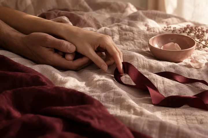 Two hands resting together on pale bedding beside a burgundy ribbon shaped into a heart.