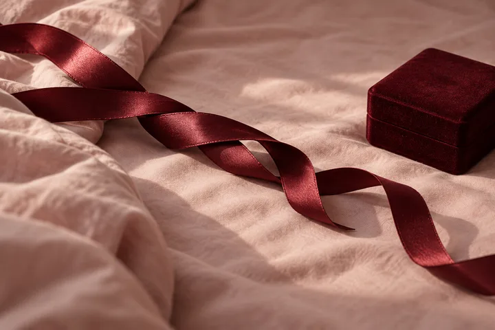 A burgundy ribbon winding across pale bedding toward a closed velvet jewelry box.