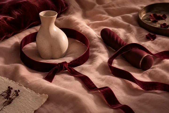 A ceramic vase and a rolled burgundy object sit inside loops of burgundy ribbon on rumpled blush fabric, with a tray of rose petals nearby.