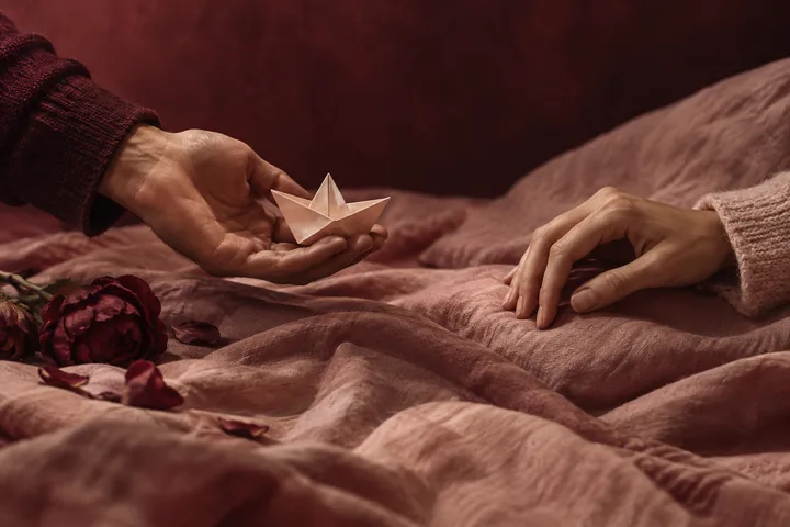 A man's hand offers a small paper boat while a woman reaches toward it without closing the space between them.
