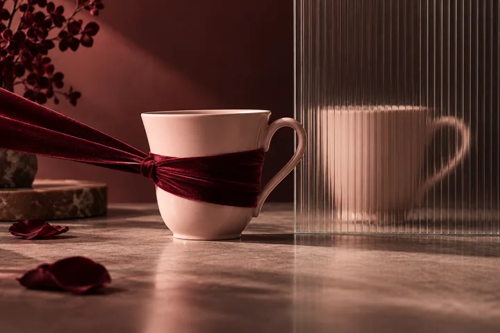 A pale cup wrapped with burgundy ribbon sits beside a ribbed glass panel that reflects another cup.