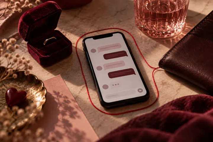 A phone with chat bubbles lies inside a loop of red cord beside an open ring box, a glass, a dark wallet, and a small heart-shaped object.