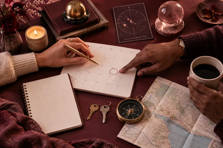Overhead burgundy editorial still life of two people planning a move together with a calendar review date, keys, compass, map, and an illustrative astrology card.