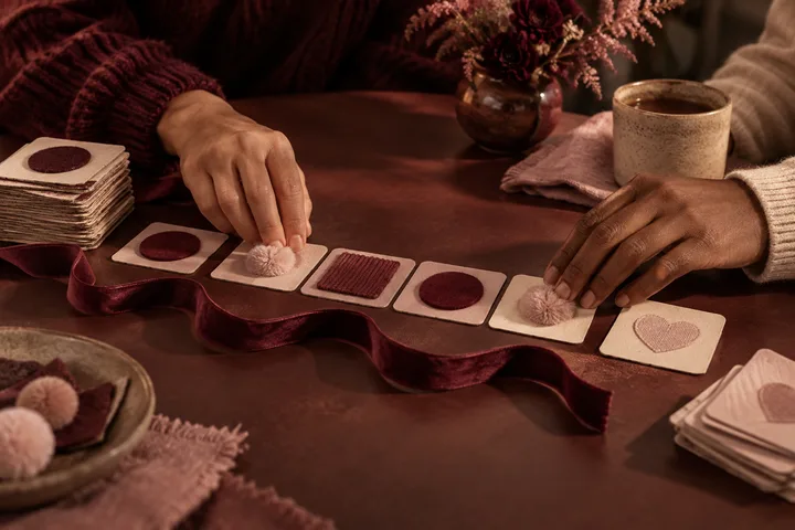 Two pairs of hands arrange burgundy and blush tactile cards with circles, fabric, and pom-poms across a table, suggesting different ways of communicating.